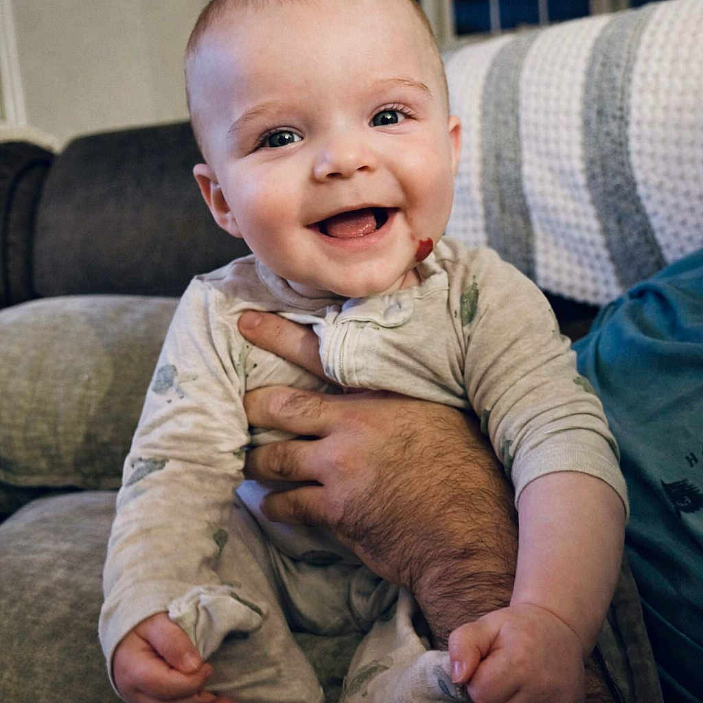 Fionn joined the competition — help win amazing prizes! baby, smiling, infant, couch, hand, adult_arm, onesie, happy, indoor, face, child, cozy, blanket, living_room, portrait, cute, skin, sitting, expression, comfort