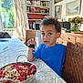 child, blue_shirt, table, bowl, red_currants, white_currants, strawberries, fruit, indoor, dining_room, chair, bookshelf, books, paintings, flower_vase, wooden_furniture, curtains, person, curly_hair, gaze