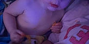Drake is registered to the contest to win money with this photo: toddler, child, snack, chips, blanket, shirtless, messy_face, indoor, dim_light, person, adult_hand, curious, blue_eyes, bed, pillow, casual, closeup, relaxed, family, resting