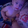 toddler, child, snack, chips, blanket, shirtless, messy_face, indoor, dim_light, person, adult_hand, curious, blue_eyes, bed, pillow, casual, closeup, relaxed, family, resting