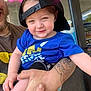 Drake is registered to the contest to win money with this photo: toddler, child, smile, hat, blue_shirt, tattoo, adult_hand, neon_sign, indoor, portrait, person, holding, casual_clothing, happy, face, cap, short_hair, skin, daylight, expression