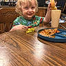 Drake is registered to the contest to win money with this photo: child, toddler, smiling, eating, food, taco, plate, table, kitchen, indoor, portrait, blonde_hair, curly_hair, green_shirt, seated, restaurant, family_meal, drink, wooden_furniture, happy