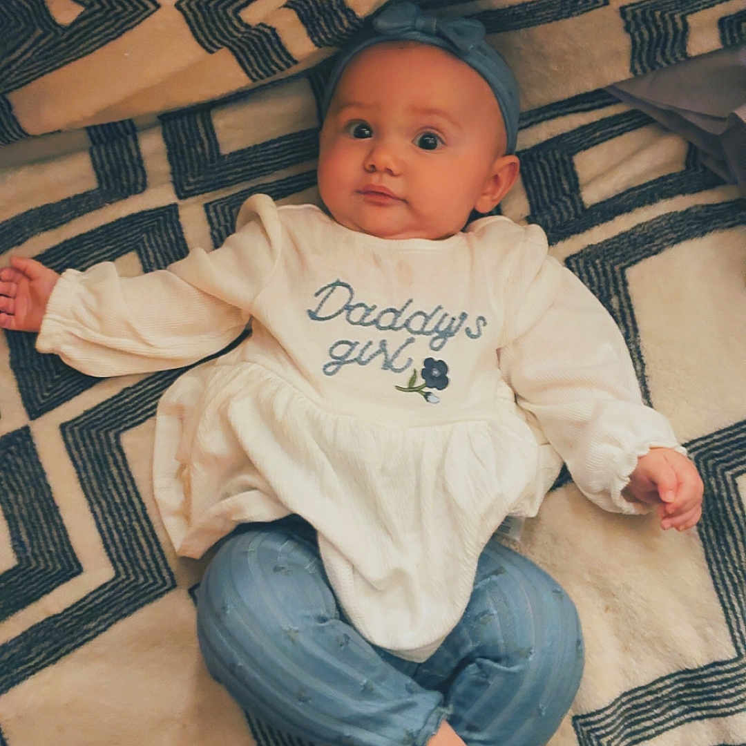 Oakley Wheeler joined the competition — help win amazing prizes! baby, child, infant, portrait, lying_down, indoors, blanket, geometric_pattern, headband, white_outfit, jeans, bare_feet, wide_eyes, curious, cute, smiling, text_on_clothing, fabric, cozy, photography