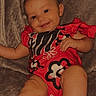 Oakley Wheeler is registered to the contest to win money with this photo: baby, child, infant, smiling, portrait, seated, indoor, chair, cushion, red_clothing, patterned_outfit, floral_pattern, face, eyes, arms, legs, feet, happy, closeup, photograph
