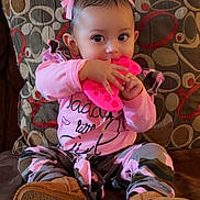Aurora is registered to the contest to win money with this photo: baby, child, girl, pink_bow, pink_clothing, camo_pants, brown_boots, toy, couch, cushion, patterned_cushion, indoor, smiling, sitting, cute, portrait, infant, hand, face, person