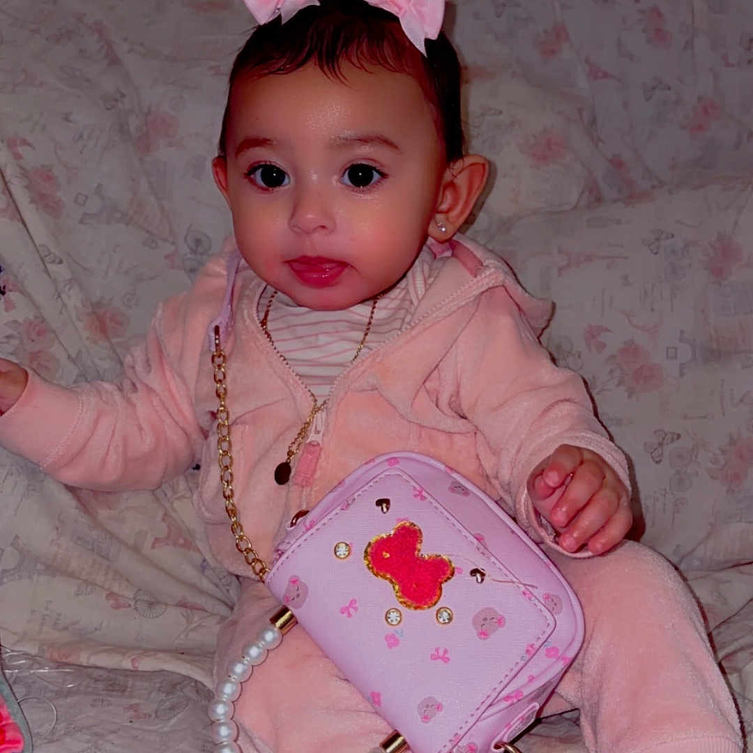 Aurora is registered to the contest to win money with this photo: toddler, child, pink_clothing, purse, bows, sitting, indoor, sofa, couch, floral_pattern, footwear, cute, accessories, young_child, portrait, person, home, decor, face, happy
