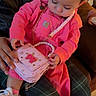 baby, child, pink, bow, purse, overalls, sneakers, person, hand, jewelry, indoor, seated, plaid_pants, jacket, curly_hair, cute, accessory, lap, focus, exploring