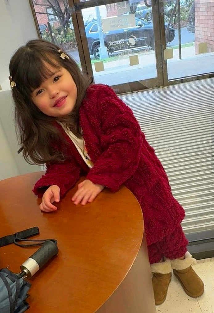Isabella Bermúdez is registered to the contest to win money with this photo: child, girl, smiling, indoor, table, wood, coat, red_clothing, boots, hair_clips, long_hair, window, glass_door, floor, casual_wear, happy, young, person, leaning, warm_clothes