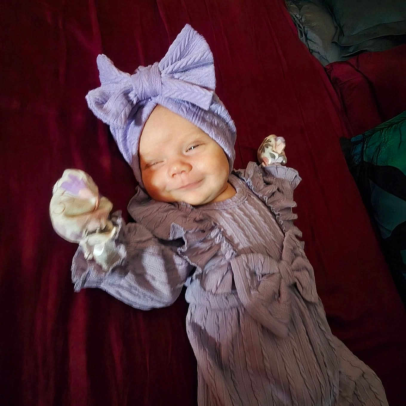 Murphy-Dean joined the competition — help win amazing prizes! baby, infant, child, smiling, portrait, lying_down, hat, headwear, dress, ruffles, blanket, bed, red_blanket, purple_clothing, mittens, indoor, cute, happy, facial_expression, close_up