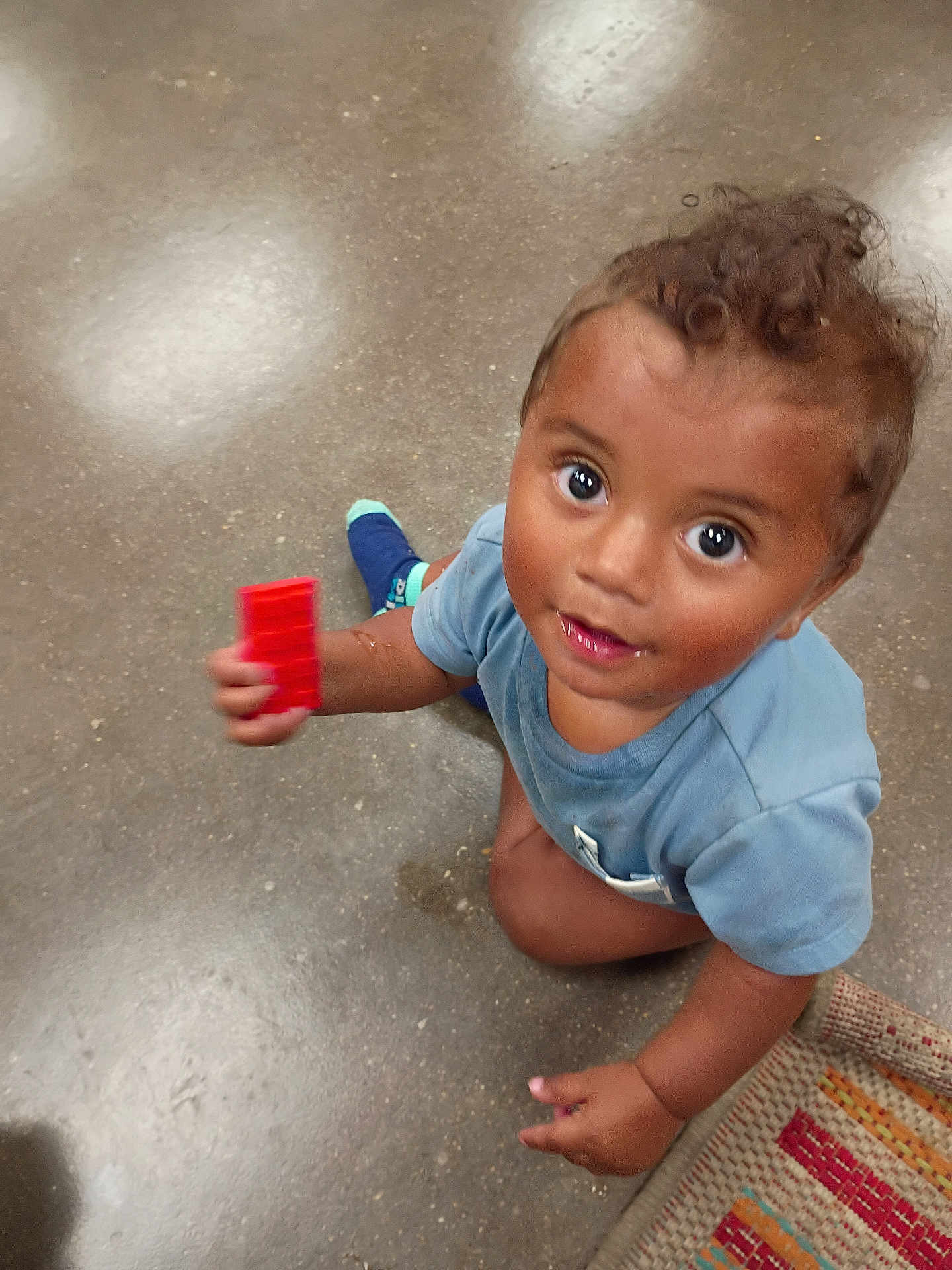 Knowledge is registered to the contest to win money with this photo: toddler, child, toy, block, floor, curly_hair, blue_shirt, indoor, play, looking_up, bright_eyes, skin, hand, kneeling, rug, colorful, casual_clothing, young_child, expression, innocence