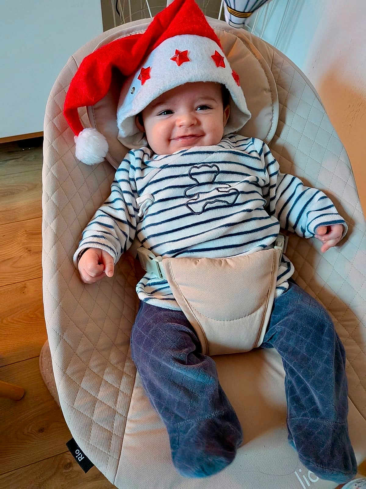 Lucas participe au concours pour gagner de l'argent avec cette photo : baby, santa_hat, striped_shirt, baby_seat, cushion, smile, indoor, wood_floor, pants, clothing, person, happy, festive, infant, cute, headwear, seatbelt, comfort, portrait, holiday