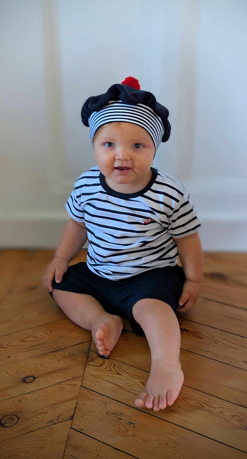 Ayden participe au concours pour gagner de l'argent avec cette photo : toddler, child, striped_shirt, beret, hat, red_pom_pom, blue_eyes, sitting, wooden_floor, barefoot, indoor, curious, smiling, person, flooring, casual_clothing, young_child, portrait, headwear, cute
