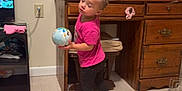 Ariyah is registered to the contest to win money with this photo: toddler, child, barefoot, pink_shirt, black_pants, toy_car, globe, wooden_desk, clutter, books, floor, indoor, curious, home, furniture, plastic_bottle, sunglasses, play, young_child, person
