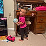 toddler, child, barefoot, pink_shirt, black_pants, toy_car, globe, wooden_desk, clutter, books, floor, indoor, curious, home, furniture, plastic_bottle, sunglasses, play, young_child, person