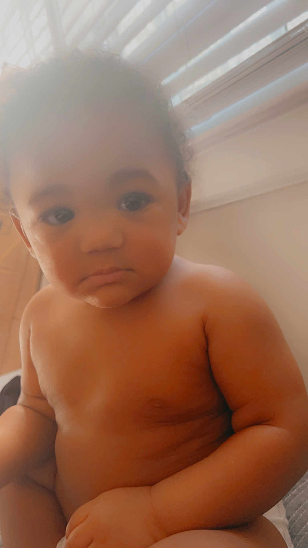 Zorian is registered to the contest to win money with this photo: baby, child, indoor, natural_light, window, blinds, skin, portrait, sitting, serious_expression, closeup, infant, curly_hair, soft_light, warm_tone, hands, face, young_child, human, room