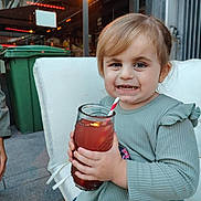 Lola a rejoint le concours — aidez-le/la à gagner de superbes lots ! toddler, child, drink, glass, straw, smile, outdoor, cushion, chair, green_trash_bin, light_hair, long_sleeve, patterned_pants, happy, person, daylight, building, concrete, hand, casual_clothing