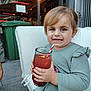 toddler, child, drink, glass, straw, smile, outdoor, cushion, chair, green_trash_bin, light_hair, long_sleeve, patterned_pants, happy, person, daylight, building, concrete, hand, casual_clothing