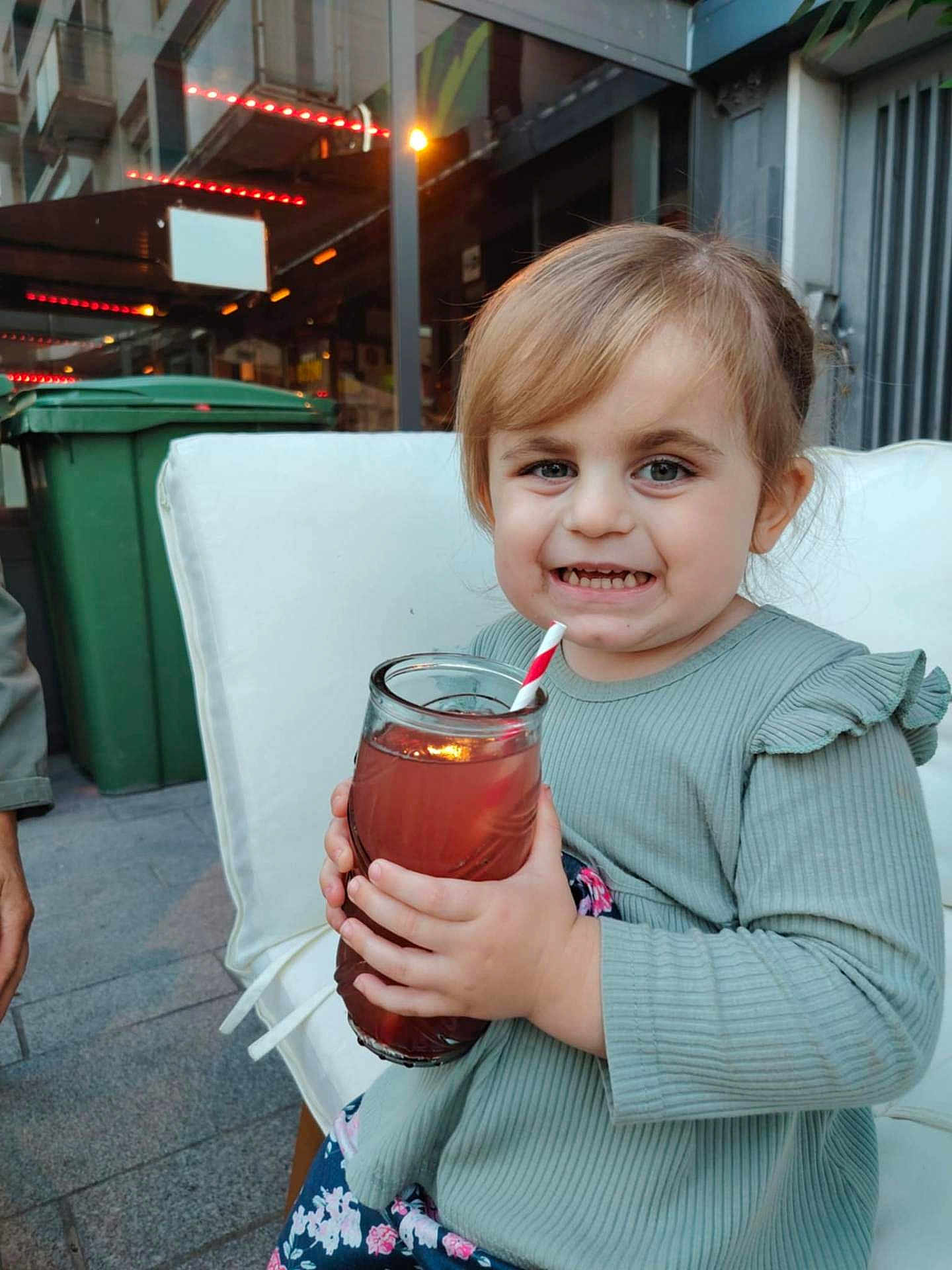 Lola a rejoint le concours — aidez-le/la à gagner de superbes lots ! toddler, child, drink, glass, straw, smile, outdoor, cushion, chair, green_trash_bin, light_hair, long_sleeve, patterned_pants, happy, person, daylight, building, concrete, hand, casual_clothing