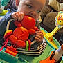 activity_center, baby, chewing, child, closeup, colorful, couch, curious, cushion, face, furniture, hand, home, indoor, infant, orange_toy, person, play, sitting, toy