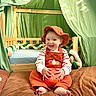 toddler, child, pumpkin, orange_overalls, hat, barefoot, brown_blanket, wooden_crib, green_fabric, tent, smiling, happy, indoors, cute, baby, playful, fall_theme, cozy, person, portrait