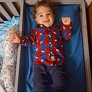 Calvin is registered to the contest to win money with this photo: toddler, crib, child, smile, happy, clothing, shirt, plaid, dog_print, pillow, bed, blanket, wooden_wall, indoor, person, furniture, baby, socks, portrait, laying_down
