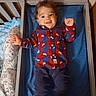 toddler, crib, child, smile, happy, clothing, shirt, plaid, dog_print, pillow, bed, blanket, wooden_wall, indoor, person, furniture, baby, socks, portrait, laying_down