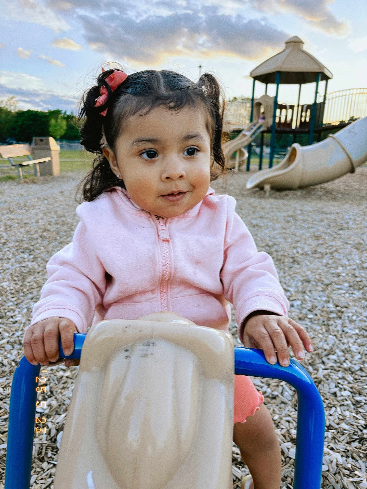 Marisol joined the competition — help win amazing prizes! toddler, child, playground, outdoor, slide, play_equipment, pink_jacket, pigtails, hair_bows, wood_chips, bench, gazebo, fence, daylight, sky, clouds, person, young_child, holding, playtime