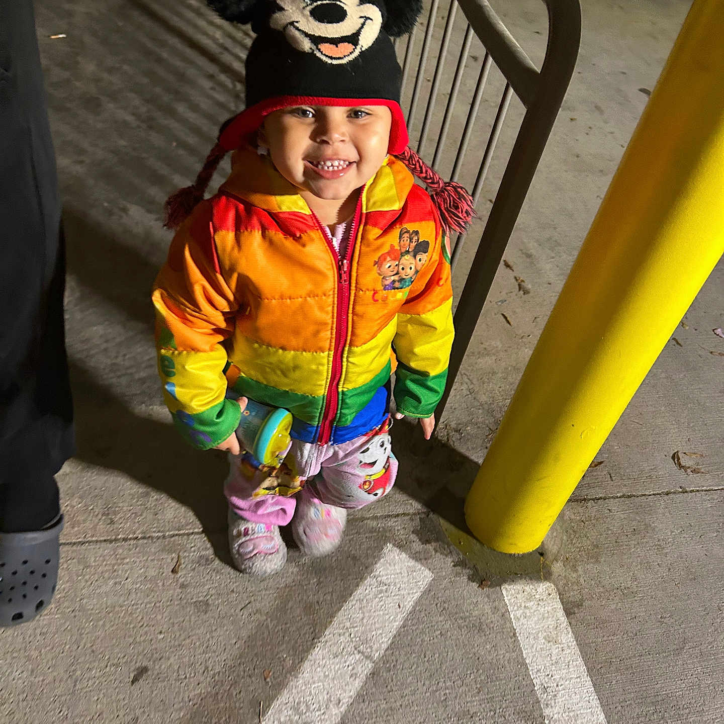 Kadayah joined the competition — help win amazing prizes! child, colorful_clothing, concrete, cute, footwear, happy, hat, night, outdoor, person, playful, railing, rainbow_jacket, sidewalk, smiling, standing, toddler, toy, warm_clothing, yellow_pole
