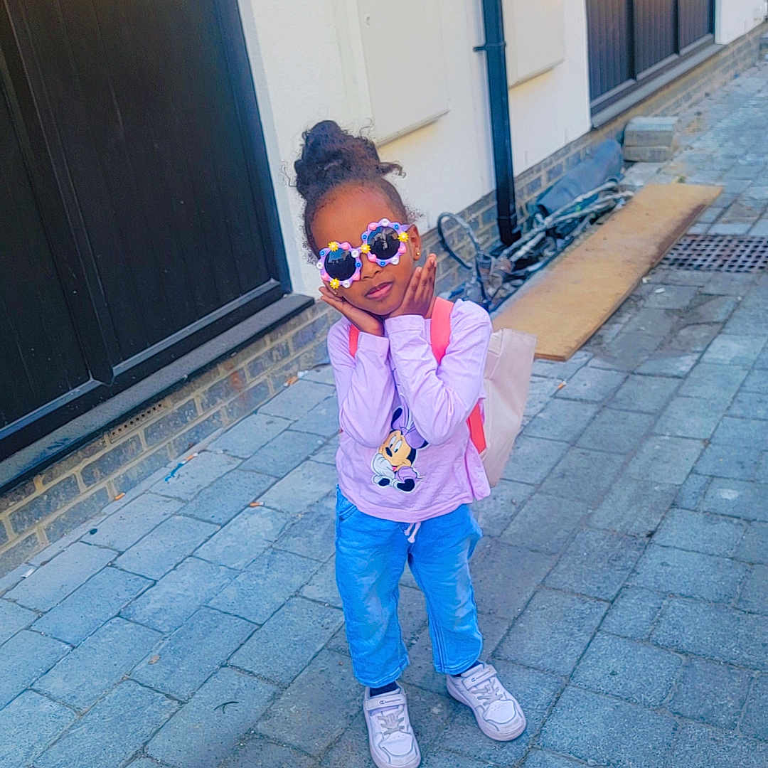 Amina joined the competition — help win amazing prizes! backpack, blue_pants, building, casual_clothing, child, cobblestone_street, curly_hair, door, flower_sunglasses, minnie_mouse, outdoor, person, pink_shirt, playful_pose, sidewalk, sunglasses, tote_bag, urban, white_sneakers, window