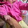 baby, child, infant, portrait, pink_jacket, yellow_bow, winter_clothing, striped_pants, sitting, being_held, adult, parent, face, eyes, smiling, indoor, close_up, colorful_clothing, family, photograph