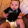 baby, infant, diaper, black_shirt, bed, pillow, patterned_pillow, lying_down, curious, wide_eyes, chubby, hands, legs, indoor, person, cute, young_child, soft_lighting, skin, face