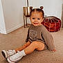 carpet, casual, child, cute, earrings, floor, footwear, hair_buns, happy, home, indoor, person, plush_pouf, portrait, sitting, smile, sweater, toddler, white_sneakers, young_child