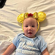 Phoenix is registered to the contest to win money with this photo: baby, infant, child, onesie, headband, yellow, red_flower, bed, white_bed_sheet, smiling, blue_clothing, face, cute, lying_down, indoor, person, portrait, young_child, happy, soft_lighting