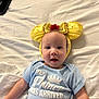 baby, infant, child, onesie, headband, yellow, red_flower, bed, white_bed_sheet, smiling, blue_clothing, face, cute, lying_down, indoor, person, portrait, young_child, happy, soft_lighting