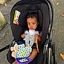 baby, car_seat, plush_toy, outdoor, pavement, autumn_leaves, baby_clothing, child, seat_belt, toy, person, feet, black_car_seat, infant, sitting, sun_pattern, colorful_toy, handle, safety, young_child