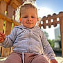child, toddler, curly_hair, blonde, gray_jacket, pink_pants, outdoor, wooden_structure, sunlight, smile, face, portrait, playground, daylight, casual_clothing, baby, young_child, happy, person, nature