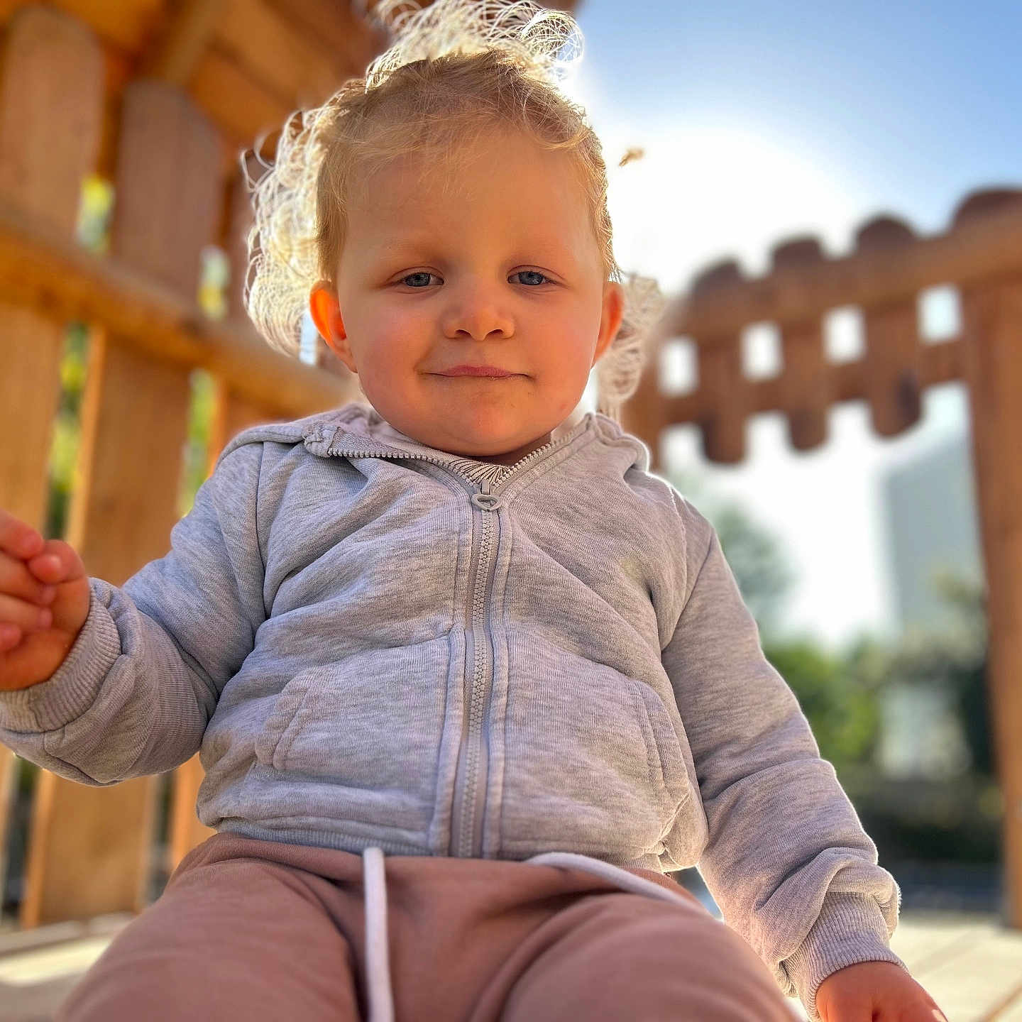 Inaya participe au concours pour gagner de l'argent avec cette photo : child, toddler, curly_hair, blonde, gray_jacket, pink_pants, outdoor, wooden_structure, sunlight, smile, face, portrait, playground, daylight, casual_clothing, baby, young_child, happy, person, nature