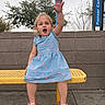 child, toddler, girl, blue_dress, pink_hearts, yellow_bench, waving_hand, outdoor, sidewalk, brick_wall, trees, cloudy_sky, sandals, happy, smiling, person, casual_clothing, playful, daytime, portrait