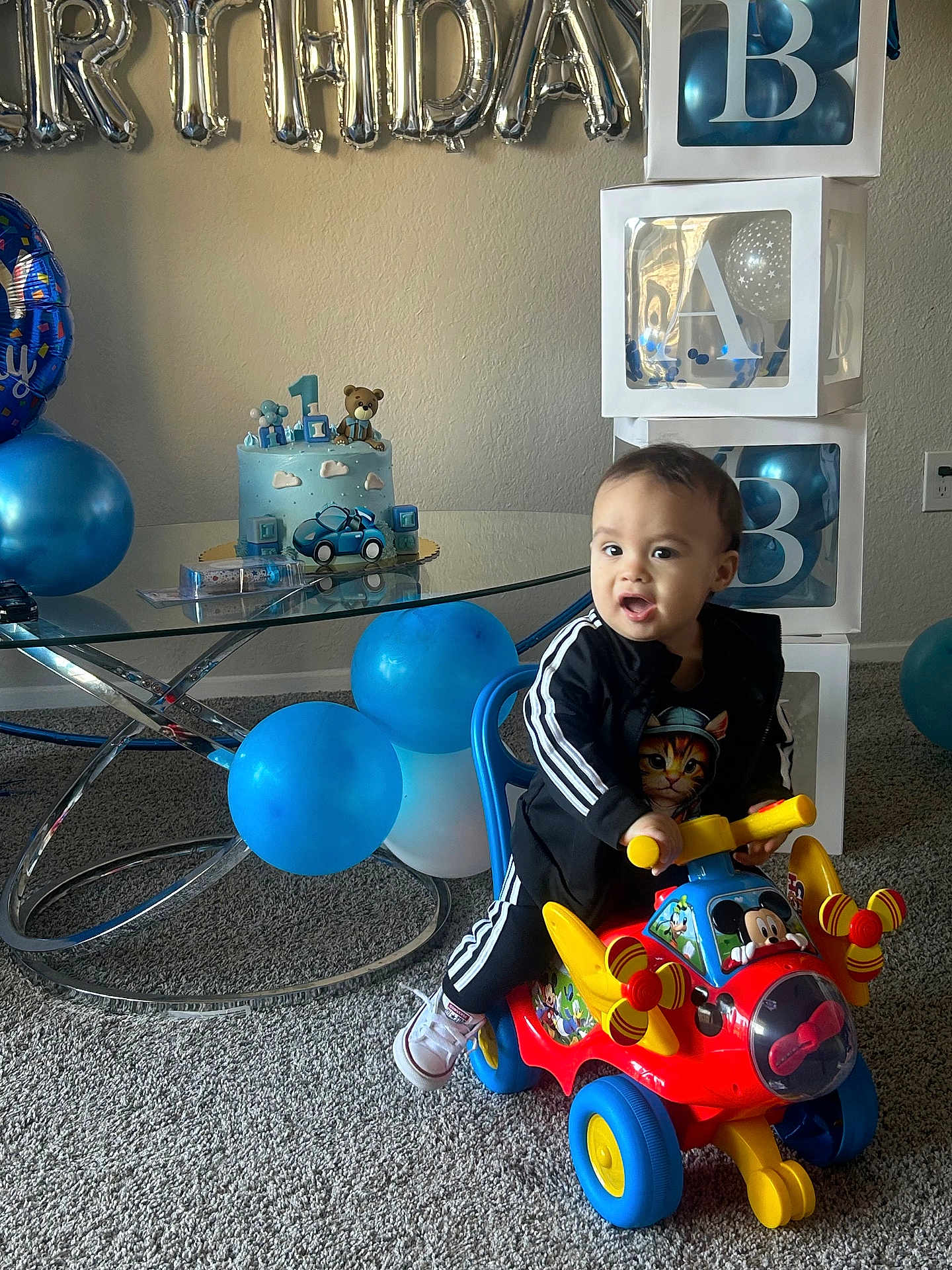 Jean Rodriguez is registered to the contest to win money with this photo: toddler, child, toy, ride_on_toy, birthday_cake, balloons, party, celebration, indoor, carpet, table, alphabet_blocks, mickey_mouse, black_tracksuit, smiling, face, person, decorations, blue, playful