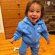 Santiago is registered to the contest to win money with this photo: child, toddler, smiling, happy, standing, portrait, blue_tracksuit, hoodie, sneakers, curly_hair, indoors, wooden_floor, dresser, fireplace, home, furniture, full_body, looking_at_camera, joyful, photograph