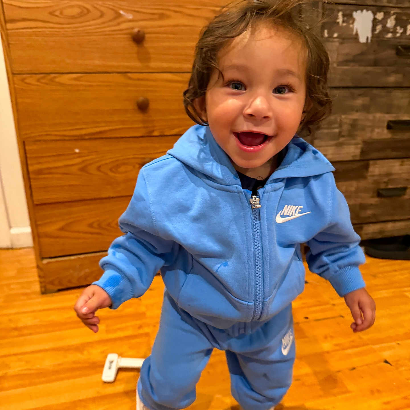 Santiago is registered to the contest to win money with this photo: child, toddler, smiling, happy, standing, portrait, blue_tracksuit, hoodie, sneakers, curly_hair, indoors, wooden_floor, dresser, fireplace, home, furniture, full_body, looking_at_camera, joyful, photograph