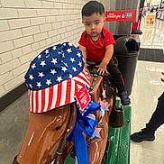 Leo joined the competition — help win amazing prizes! child, toddler, boy, portrait, sitting, mechanical_horse, ride, arcade, supermarket, indoor, red_shirt, black_pants, patriotic, american_flag, stars_and_stripes, brick_wall, store, shopping, serious_expression, horse