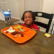 Sy is registered to the contest to win money with this photo: toddler, child, smiling, braided_hair, dining_table, orange_plate, chicken_nuggets, corn, ranch_dressing, sippy_cup, high_chair, fork, food, indoor, happy, meal, wooden_table, black_chair, person, cute