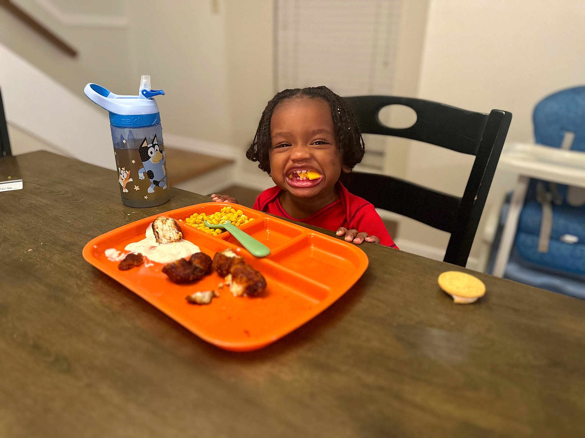 Sy is registered to the contest to win money with this photo: toddler, child, smiling, braided_hair, dining_table, orange_plate, chicken_nuggets, corn, ranch_dressing, sippy_cup, high_chair, fork, food, indoor, happy, meal, wooden_table, black_chair, person, cute