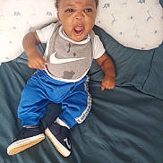 Je'Saiah joined the competition — help win amazing prizes! baby, child, expressive_face, gray_bib, blue_pants, sneakers, sitting, blanket, pillow, animal_pattern, indoors, cute, clothing, footwear, person, young_child, happy, playful, portrait, home