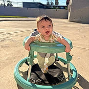 Carlos is registered to the contest to win money with this photo: baby, walker, outdoor, sunlight, concrete, palm_tree, happy, child, infant, play, sunny, casual_clothing, footwear, expression, daylight, shadow, background, person, head, smile