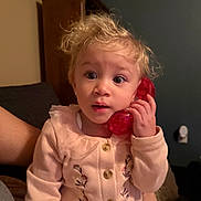 Isabella is registered to the contest to win money with this photo: background, buttoned_sweater, child, curious, curly_hair, cute, earrings, expression, face, furniture, hand, home, indoor, person, pink_clothing, sitting, toddler, toy_phone, warm_lighting, young_child