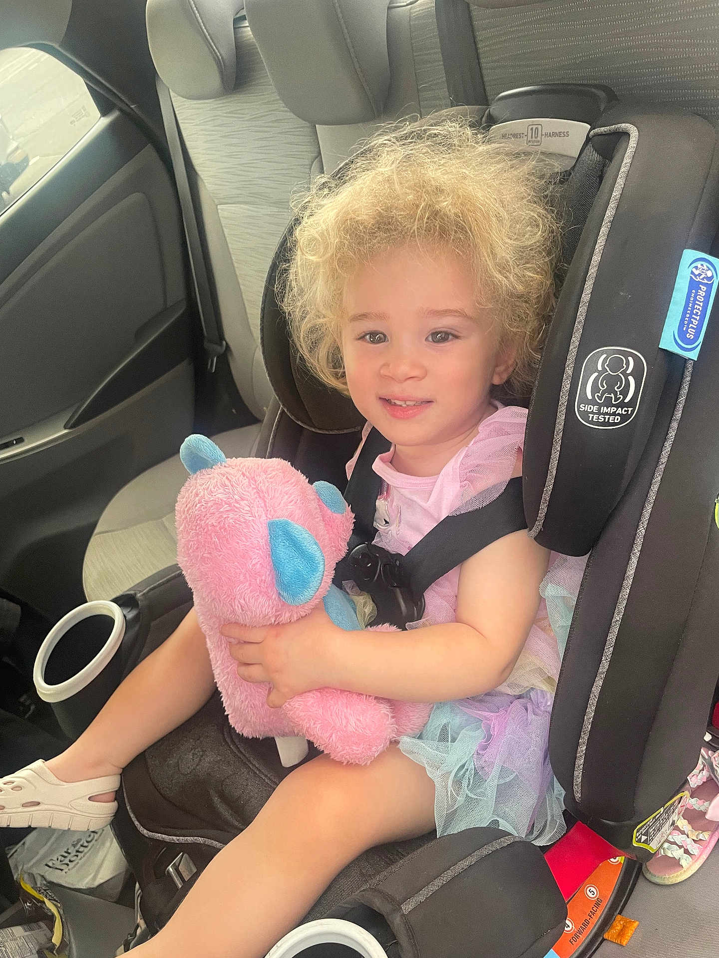Isabella joined the competition — help win amazing prizes! toddler, child, car_seat, stuffed_animal, toy, curly_hair, smile, pink, blue, pastel_colors, dress, footwear, car_interior, seatbelt, happy, sitting, young_child, safety, vehicle, cute