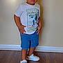 beige_wall, boy, casual_clothing, child, curious_expression, dinosaur_print, flooring, footwear, full_body, hands_on_hips, indoor, looking_away, portrait, short_hair, shorts, standing, tshirt, white_shoes, wooden_floor, young_child