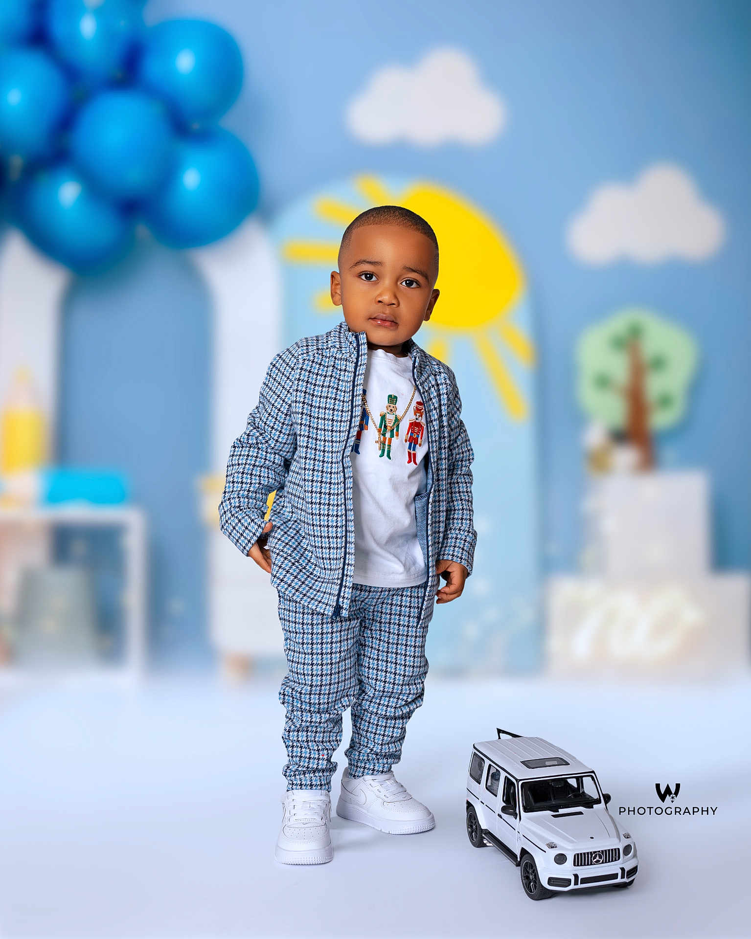 Josiah joined the competition — help win amazing prizes! toddler, child, blue_outfit, checkered_clothing, white_shoes, toy_car, balloons, sun, clouds, tree, indoor, portrait, fashion, cute, standing, playful_background, photography, smiling, young_child, casual